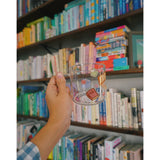 A glass mug with a design of colorful books printed all around. The mug is held up by a hand in front of a bookcase full of colorful books. From Books Unbound.