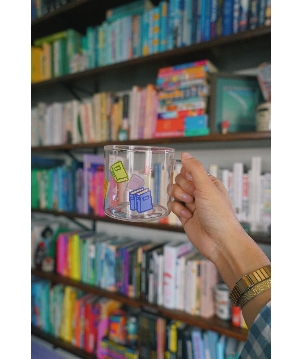 A glass mug with a design of colorful books printed all around. The mug is held up by a hand in front of a bookcase full of colorful books. From Books Unbound.