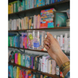 A glass mug with a design of colorful books printed all around. The mug is held up by a hand in front of a bookcase full of colorful books. From Books Unbound.