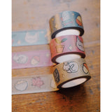 Three washi tapes of different colorways taped across the top of a table. The patterns on the washi tape include a owl, books, plants, mugs, cats, and more. The washi tapes come in a set of blue, purple, and brown colors. From Books Unbound.