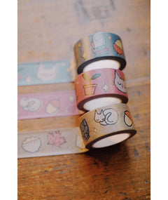 Three washi tapes of different colorways taped across the top of a table. The patterns on the washi tape include a owl, books, plants, mugs, cats, and more. The washi tapes come in a set of blue, purple, and brown colors. From Books Unbound.