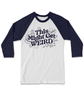 A white baseball tee with dark blue arms and fun, curvy dark blue text that reads "This Might Get WEIRD" across the chest. Around the text are illustrated outlines of objects like hot dogs, cats, stars and planets.