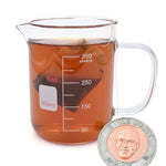 A glass beaker with a mug handle full of brown tea and a tea bag. On the side of the mug is the NileRed logo and a square that says 400mL and shows a scale of 350mL. To the side is a coin with an image of Nigel from NileRed and the text "2 Nile Bucks; 2025" with a diamond fractal pattern.