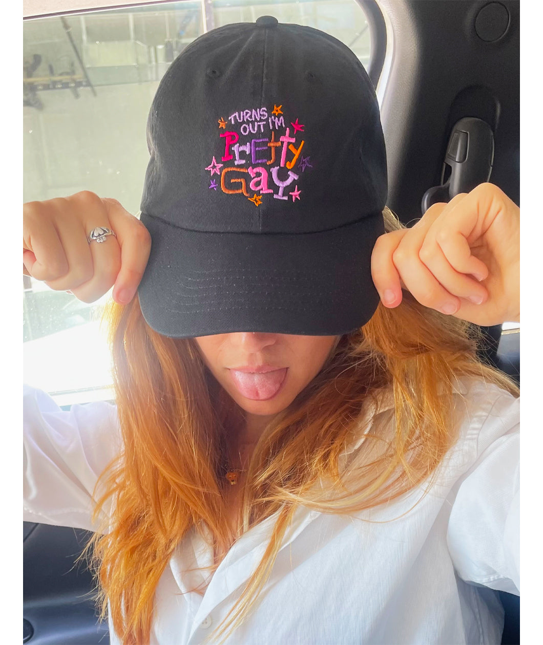 A photo of Catherine McCafferty posing, wearing a black cap. The cap reads "Turns Out I'm Pretty Gay" and has little star illustrations around it. From Pretty Gay.
