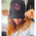 A photo of Catherine McCafferty posing, wearing a black cap. The cap reads "Turns Out I'm Pretty Gay" and has little star illustrations around it. From Pretty Gay.
