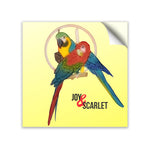Two mackaws sitting on a tan dowell. Mackaw in front is red, green, and blue. Behind is another mackaw colored yellow, green, and blue. Both are in front of small tan circle. Inside of circle are objects hanging. “Joy & Scarlet” is written below mackaws in black sans serif font. The “&” is italicized and red. The base color of sticker is yellow - from Animal Wonders