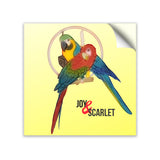 Two mackaws sitting on a tan dowell. Mackaw in front is red, green, and blue. Behind is another mackaw colored yellow, green, and blue. Both are in front of small tan circle. Inside of circle are objects hanging. “Joy & Scarlet” is written below mackaws in black sans serif font. The “&” is italicized and red. The base color of sticker is yellow - from Animal Wonders