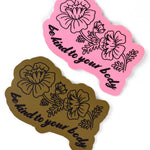 Two stickers both with a black outline of flowers with a stem attached and some leaves. Underneath, “be kind to your body” is in black cursive font below in a wavy style. One sticker has a pink base and the other a dark gold base - from Sierra Schultzzie