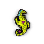 A Hank Green enamel pin of a crude dragon drawn out of scribbles. The dragon is green with a blue mane of hair and has a yellow heart and tail.
