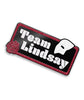 Rectangle pin with curved edges, red glitter outline and black fill. Bottom left of rectangle is a red glitter rose. Top right is half of a white glitter mask. “Team Lindsay” is inside of rectangle in white glitter serif font - from Linsay Ellis. 