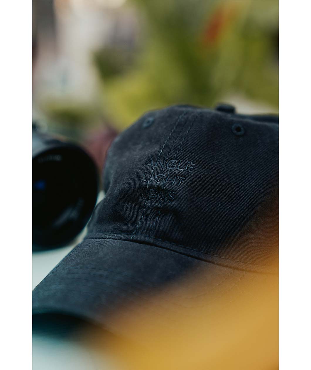 A close up of a black hat embroidered with "Angle Light Lens" sits on a table with a camera next to it.