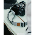 A close up of a grey camera strap attached to a camera with a brown leather square that has a "LP" etched into it for Lizzie Peirce.
