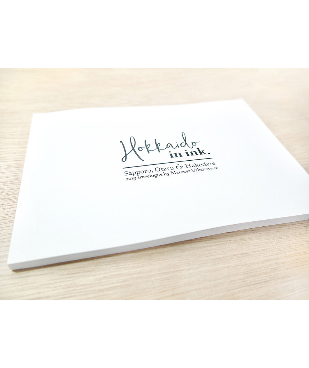 A flat, thin, white book with the words "Hokkaido in ink." above "Sopporo, Otaru & Hakodate 2019 travelogue by Mateusz Urbanowicz" in black writing. 