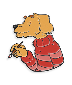 A pin shaped like the top half of a dog. The dog is a sandy color swearing a striped red sweater and holding a paintbrush. The painting dog pin from Mateusz Urbanowicz.