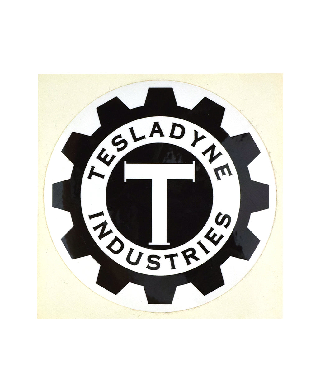 A circular white sticker with a black gear. Gear surrounds a white circle which has a black circle in the middle. A white serif “T” is in the center. “Tesladyne Industries” is in black serif font arched around the middle black circle - from Tesladyne