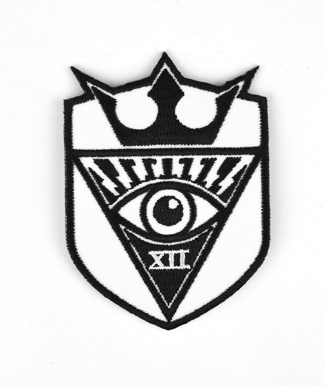 White sheild patch with a black outline. There is an upside down black triangle with a black and white eye in it with XII below in white font. A black crown is on top of the triangle - from Tesladyne