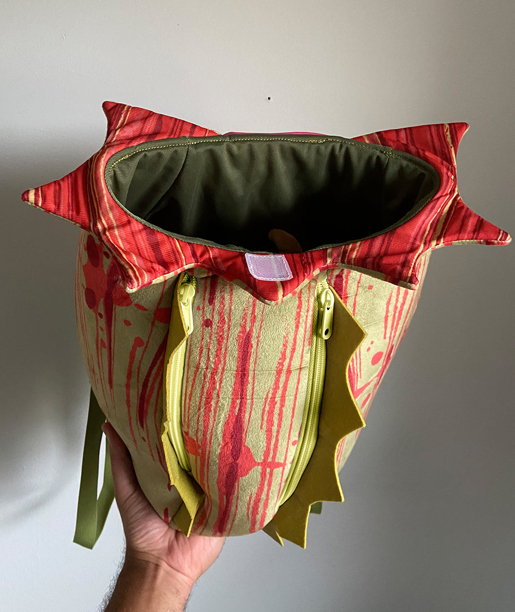 A soft, green and red backpack made to look like a pitcher plant. The top is jagged, the back has two zippers, each lined with spiky green and has solid green straps. From Tyler Thrasher.
