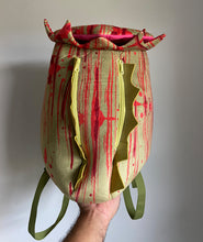 A soft, green and red backpack made to look like a pitcher plant. The top is jagged, the back has two zippers, each lined with spiky green and has solid green straps. From Tyler Thrasher.