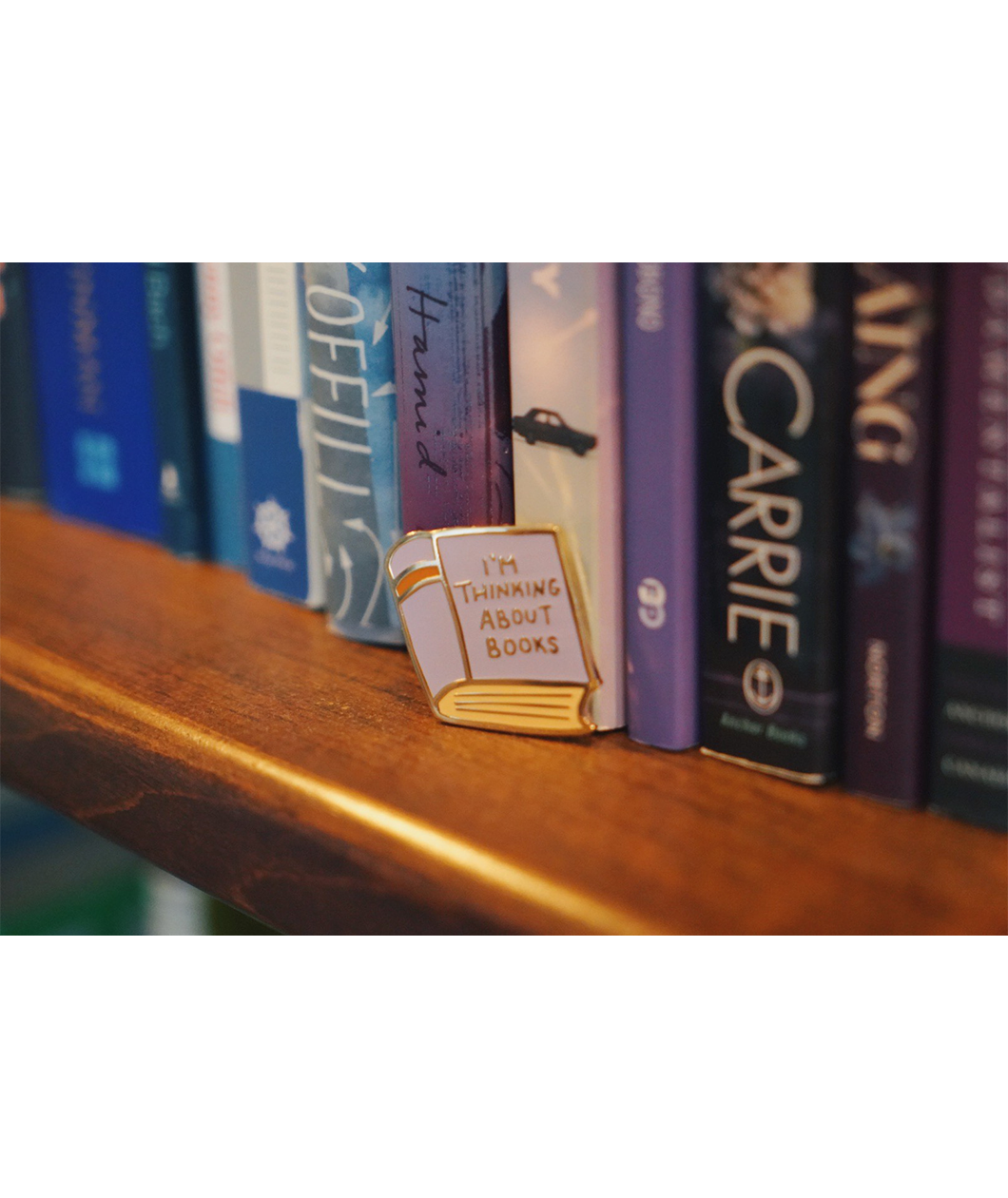 A gold and purple pin shaped like a book. The cover of the book says "I'm thinking about books" in front of books on a bookshelf. From Ariel Bissett.