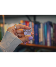 A gold and purple pin shaped like a book. The cover of the book says "I'm thinking about books" in front of books on a bookshelf. From Ariel Bissett.