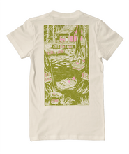 A t-shirt with a rectangular print on the back of an illustrated swamp in green and pink. The print features a house, trees, a bed and boat floating in the swamp with lily pads. Atop the bed and boat are people reading a book. Books are scattered and hidden throughout the swamp. From Ariel Bissett.