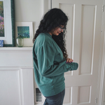 Ariel Bissett holding the left sleeve of her alpine green sweatshirt which has a small mint green outline of a book on it.