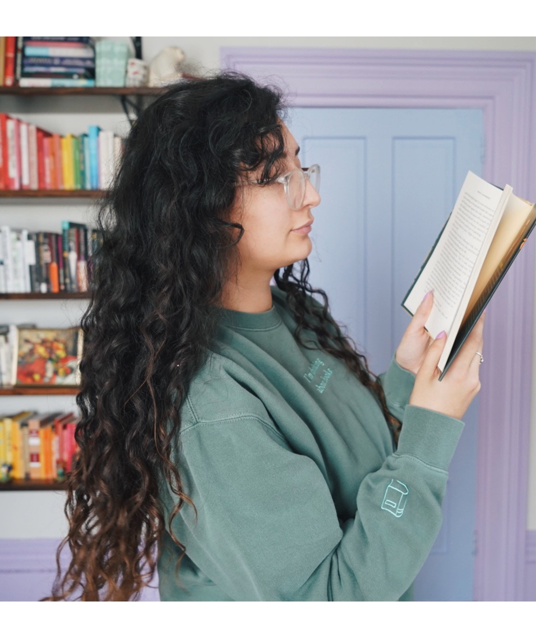 Ariel Bissett reading while wearing an alpine green sweatshirt with a mint green outline of a book on the sleeve.