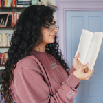 Ariel Bissett reading while wearing a dusty rose sweatshirt with a mint green outline of a book on the sleeve.