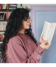 Ariel Bissett reading while wearing a dusty rose sweatshirt with a mint green outline of a book on the sleeve.