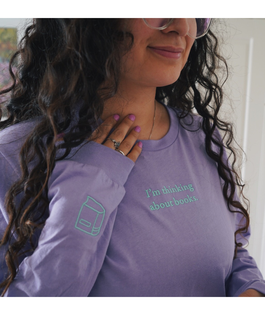 A close-up of the top half of a lavender long sleeve shirt worn by Ariel Bissett. It has the phrase "I'm thinking about books" on the upper chest and the outline of a book on the left sleeve, both in mint.