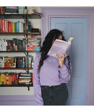 Ariel Bissett standing by a bookshelf and reading while wearing a lavender long sleeve shirt.