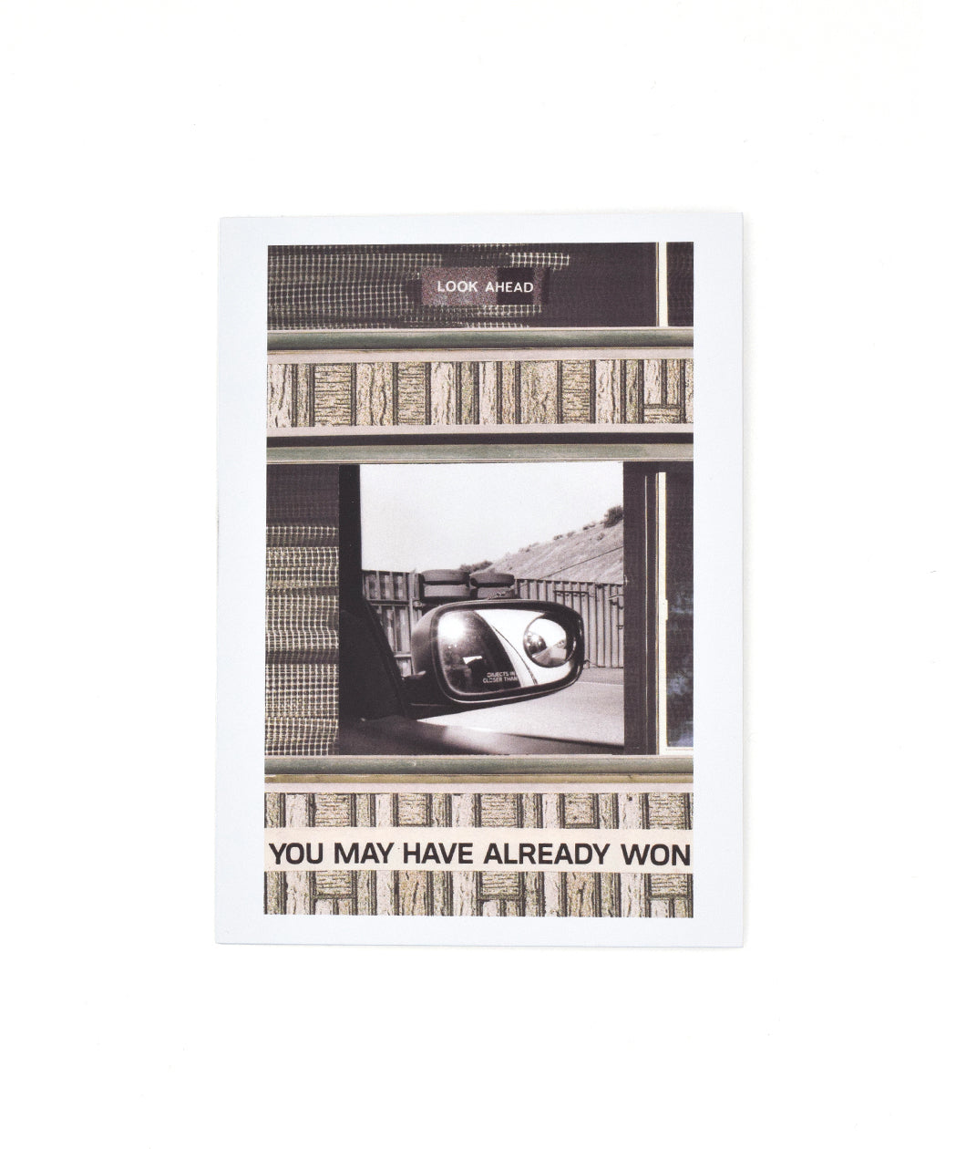 A white greeting card with a black and white image a car mirror with a fence and hill in the background. Textured designs are all around the image. “Look ahead” is in white sans serif font at the top. “You may have already won” in black sans serif font at the bottom - from the Art Assignment