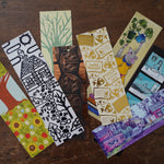 8 different illustrated, colorful bookmarks from the Books Unbound subscription laying overlapping on a table. 