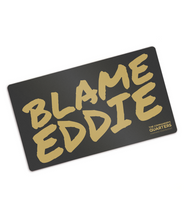 Black rectangular playmat with curved corners. “Blame Eddie” is in gold font resembling handwriting. “The commander’s quarters” is in the bottom right corner in gold sans serif font - from The Commander’s Quarters