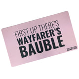 A pink rectangular playmat with curved edges. “First Up There’s Wayfarer’s Bauble” is in black sans serif font in varying weights in the center. “The Commander’s Quarter’s” is in the bottom right of the playmat - from the Commander’s Quarters