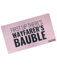 A pink rectangular playmat with curved edges. “First Up There’s Wayfarer’s Bauble” is in black sans serif font in varying weights in the center. “The Commander’s Quarter’s” is in the bottom right of the playmat - from the Commander’s Quarters
