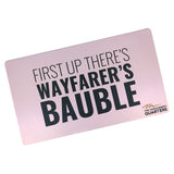 A pink rectangular playmat with curved edges. “First Up There’s Wayfarer’s Bauble” is in black sans serif font in varying weights in the center. “The Commander’s Quarter’s” is in the bottom right of the playmat with a signature above - from the Commander’s Quarters