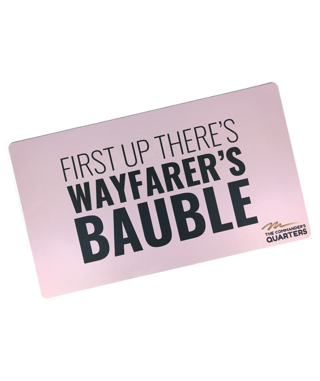 A pink rectangular playmat with curved edges. “First Up There’s Wayfarer’s Bauble” is in black sans serif font in varying weights in the center. “The Commander’s Quarter’s” is in the bottom right of the playmat with a signature above - from the Commander’s Quarters