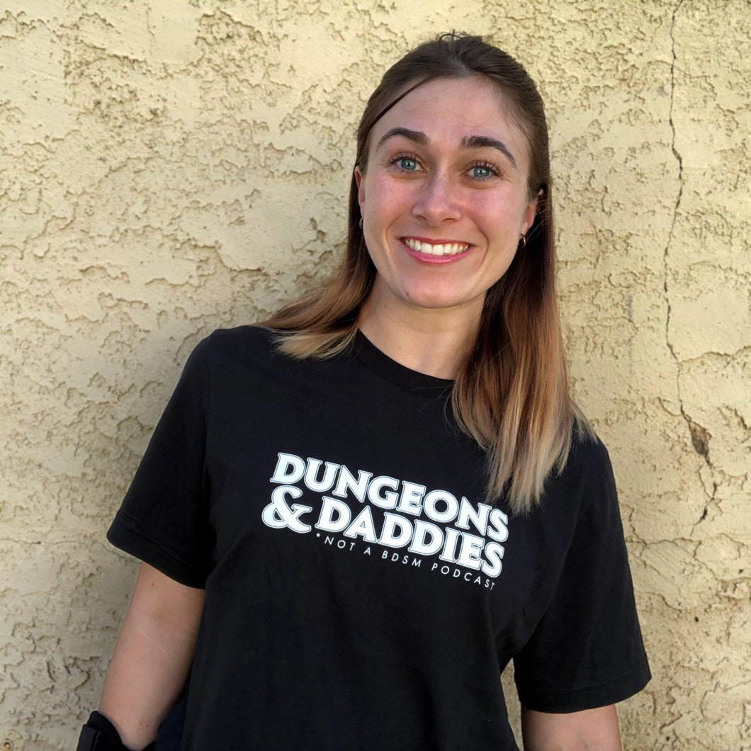 Beth May, a woman with blue eyes and blonde balyaged hair with brown roots smiling, modeling the Dungeons & Daddies logo shirt.