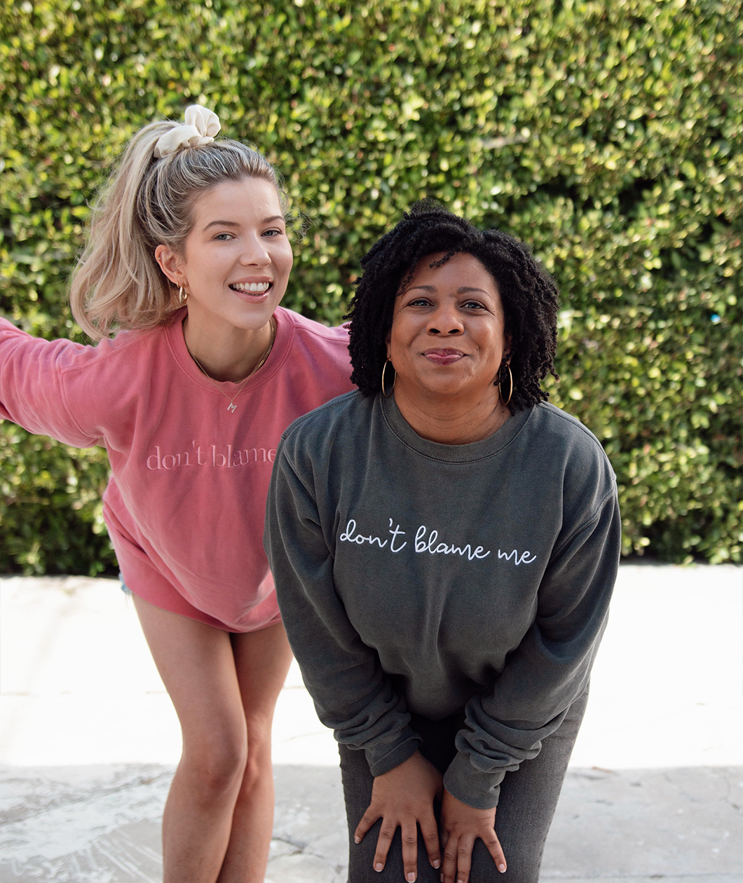 Meghan Rienks modeling a pink crewneck sweatshirt with “don’t blame me” across the chest in pink serif font and Melisa Monts modeling a gray crewneck sweatshirt with “don’t blame me” across chest in white cursive font - from Don’t Blame Me