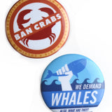 One red and white pin with a drawing of a crab with the words "ban crabs" striking through it. One blue and white pin with a drawing of a fist holding a whale and the words "We demand whales. Also, what are they?" - by Door Monster