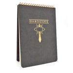 A large black sketchbook with gold spirals at the top and the word "drawtectives" with a drawing of a sword and an eye in gold in the center - by Drawfee