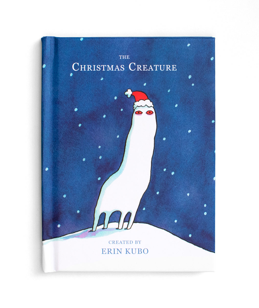 A book with a four legged white creature with a tall neck and red eyes with a Santa hat on standing in the snow against a blue background and snow falling behind. “The Christmas Creature” is at the top in white serif font - from Erin Kubo