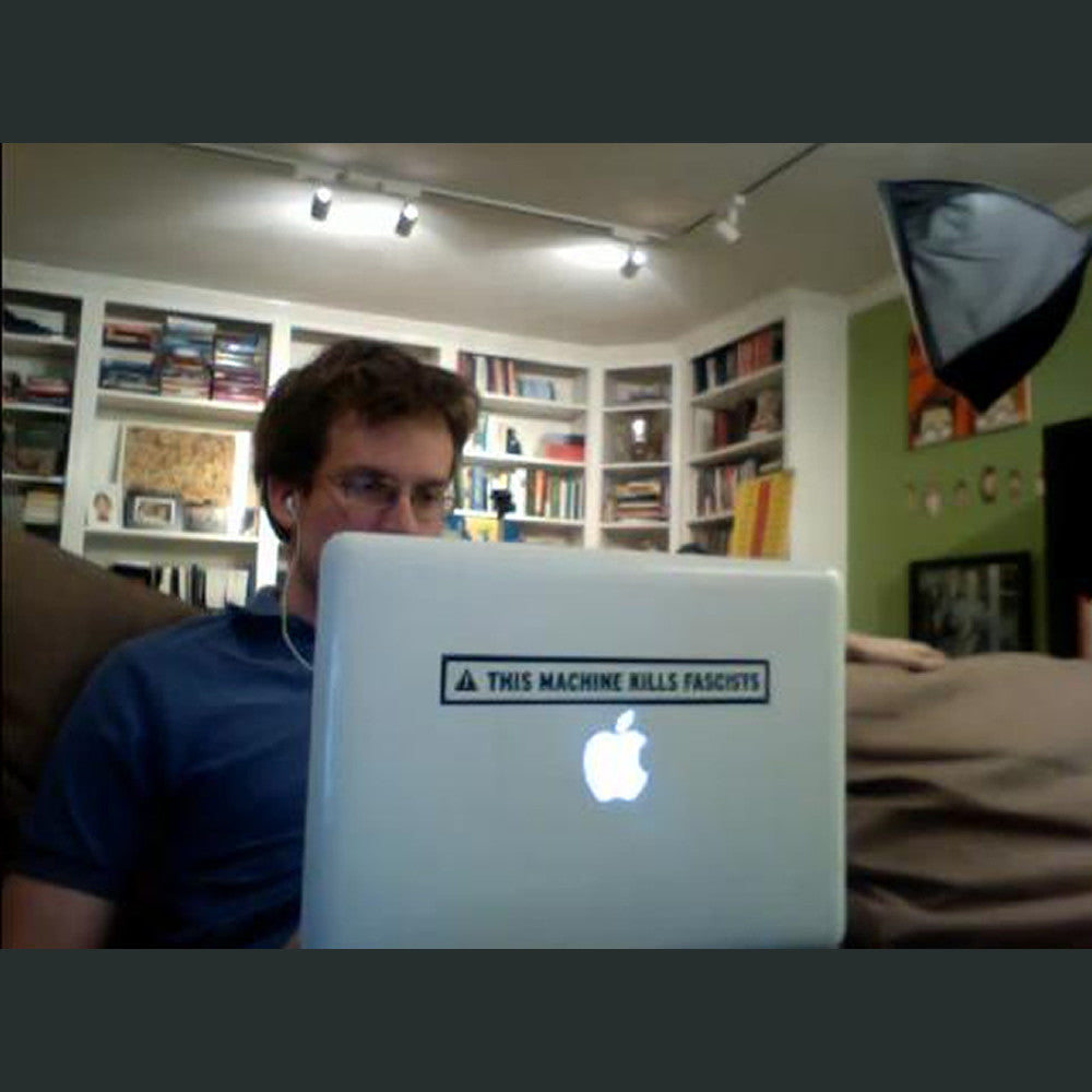 John Green sitting on a couch on his computer with the sticker on the back of his laptop - from John Green