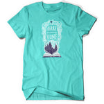 A light teal shirt with an open book with a white outline drawing with purple fill of Hogwarts. White intertwined lined surround “Harry I’m Coming Home” in white font. A purple heart is atop - from Lauren Fairweather