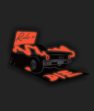 The glow in the dark form of the Helix pin from Mars Heyward. The pin shows the back of a black car with the red smoke coming out of the exhaust pipe glowing and the numbers "666" on the tail end of the car. Above the car it says "Ride or" and on the ground behind the car, it says "Die". The words are all part of the pin that is glowing red. 