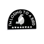 Black half circle pin with a thin silver border. “I’m losing to a bird” is arched in silver sans serif font. In the center is a silhouette drawing of a silver bird with a black eye holding four cards standing above card suits - from Lindsay Ellis