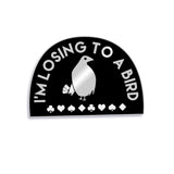 Black half circle pin with a thin silver border. “I’m losing to a bird” is arched in silver sans serif font. In the center is a silhouette drawing of a silver bird with a black eye holding four cards standing above card suits - from Lindsay Ellis