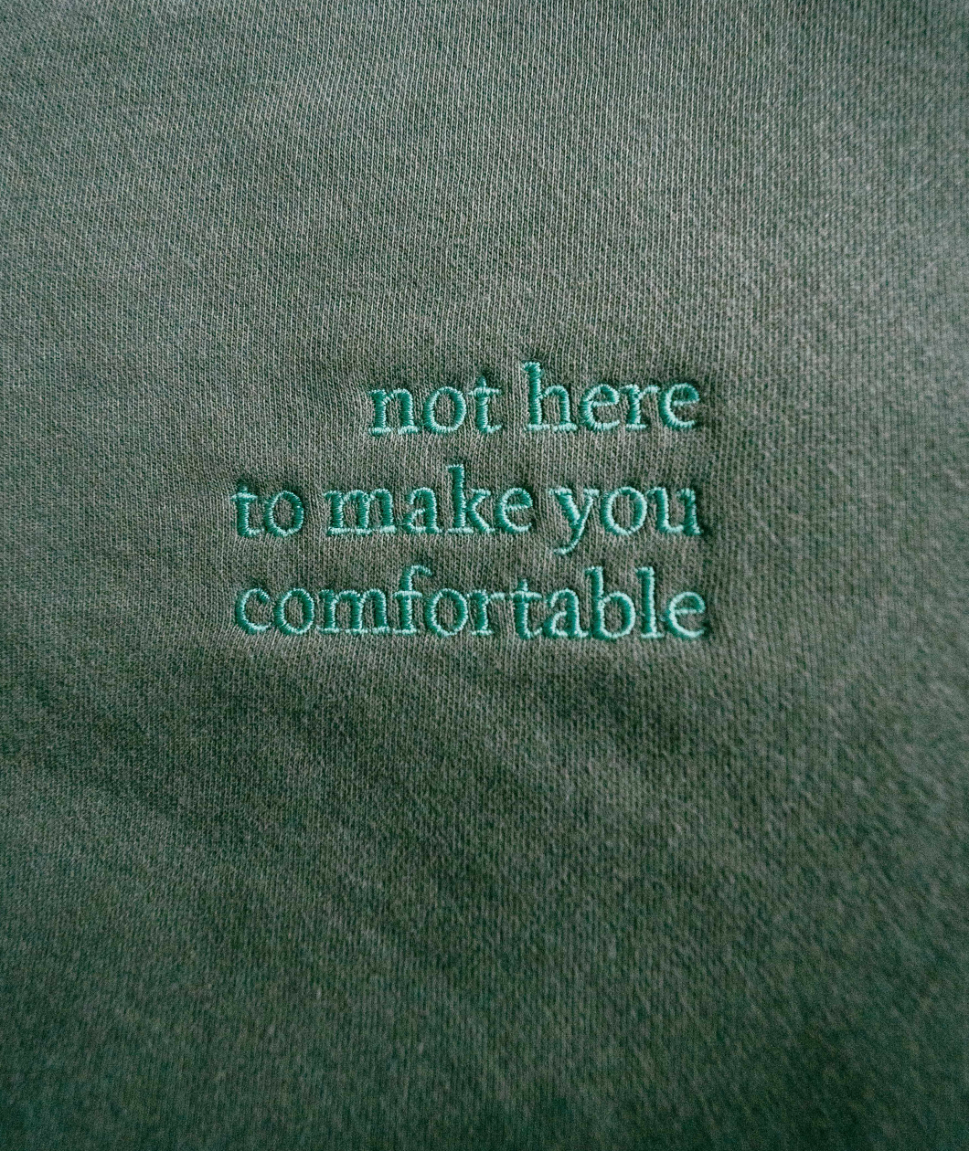 “not hear to make you comfortable” embroidered in green serif font on a green swater - from Iz Harris