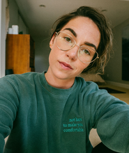 Iz Harris modeling a green crewneck sweatshirt with “not hear to make you comfortable” embroidered in green serif font at the top right of the sweater - from Iz Harris
